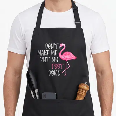 Foot awareness pink flamingo apron - Pink flamingo standing next to stacked text don't make me put my foot down — shown on a black cotton canvas apron against a white background
