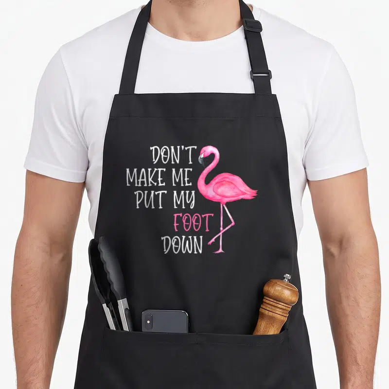 Foot awareness pink flamingo apron - Pink flamingo standing next to stacked text don't make me put my foot down — shown on a black cotton canvas apron against a white background