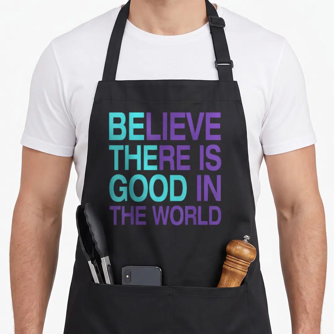 Believe motivation apron - Stacked teal and purple text reading believe there is good in the world — shown on a black cotton canvas apron against a white background