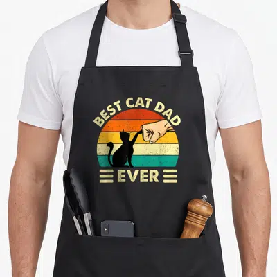 Owner kitty cat apron - Retro sun graphic with black cat fist bumping human hand and text BEST CAT DAD EVER — shown on a black cotton canvas apron against a white background