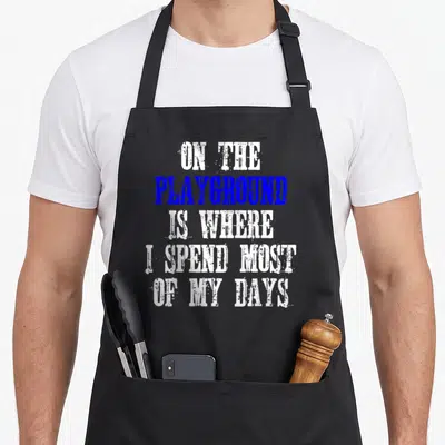 Cute most playground apron - Five-line stacked text in white and blue distressed font reading ON THE PLAYGROUND IS — shown on a black cotton canvas apron against a white background