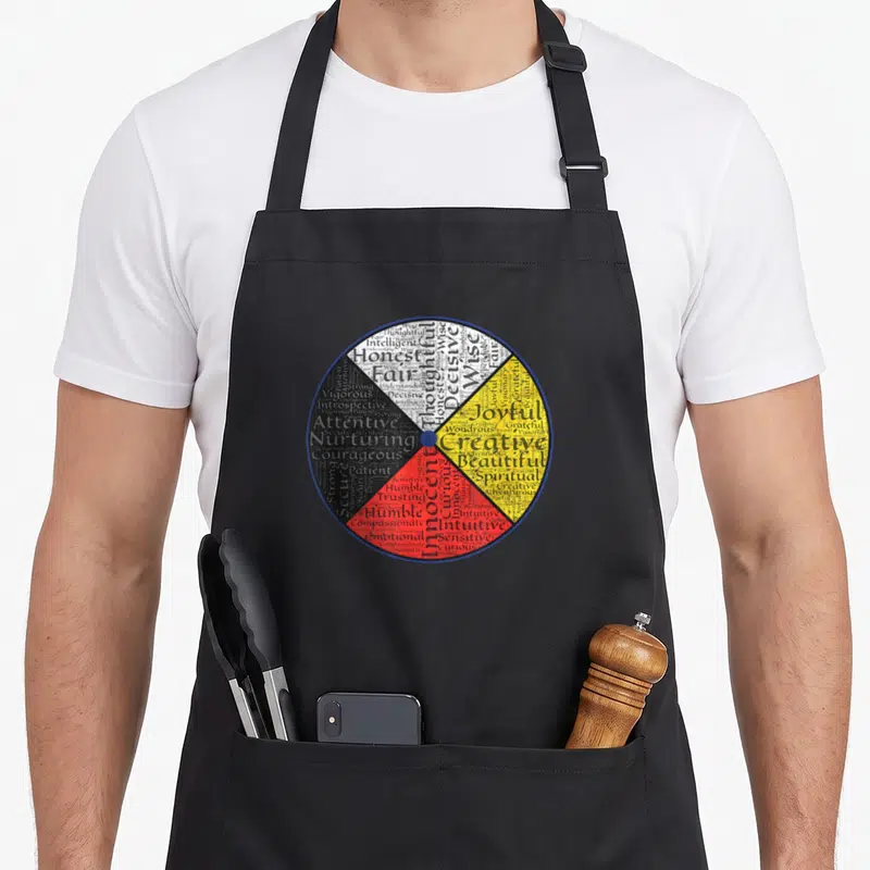 Native wheel apron - Circular typography art with virtues like Wise and Humble arranged in white, yellow, red — shown on a black cotton canvas apron against a white background