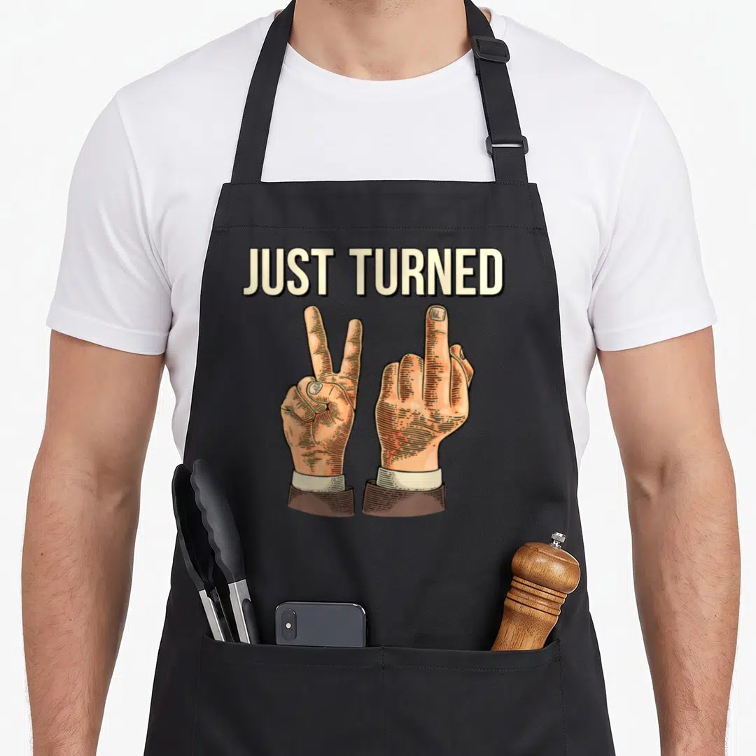 Turned birthday apron - JUST TURNED text above two cartoon hands showing a peace sign and middle finger in tan and brown — shown on a black cotton canvas apron against a white background