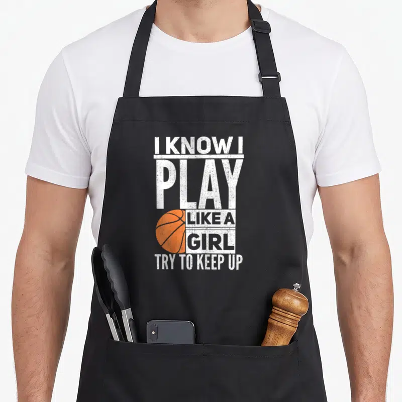 Try basketball apron - Stacked white text I KNOW I PLAY LIKE A GIRL TRY TO KEEP UP beside orange basketball — shown on a black cotton canvas apron against a white background
