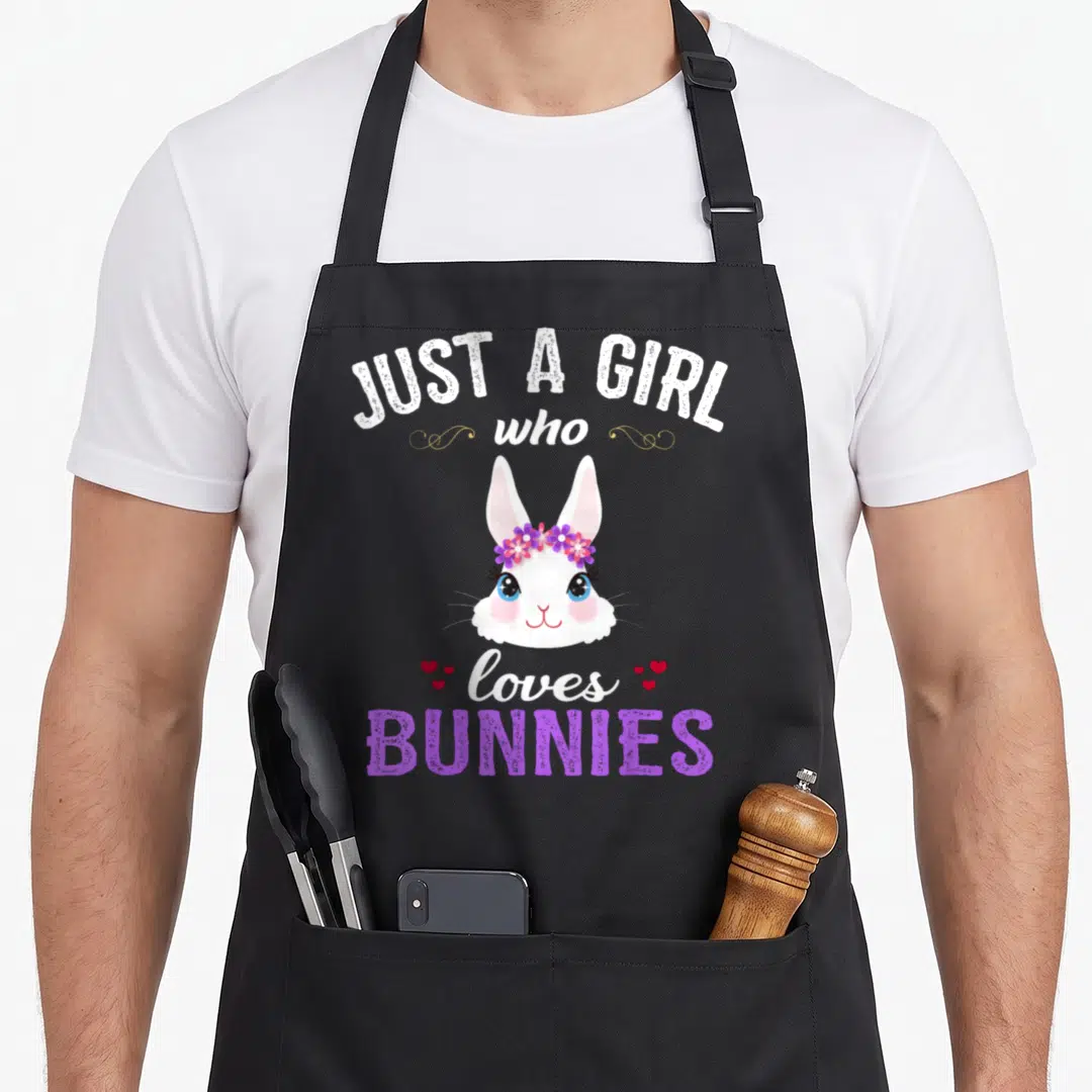 Cute bunny apron - Cartoon white bunny face with purple flower crown and text JUST A GIRL who loves BUNNIES surrounded — shown on a black cotton canvas apron against a white background