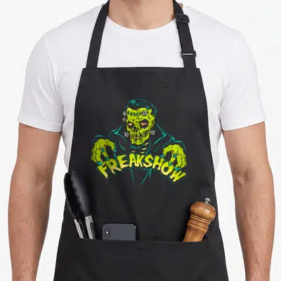 Zombie horror apron - Cartoon zombie in teal coat with lime green skin and FREAKSHOW text overlapping its hands — shown on a black cotton canvas apron against a white background