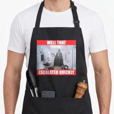 Well general apron - Darth Vader in white corridor with red banners reading Well That Escalated Quickly — shown on a black cotton canvas apron against a white background