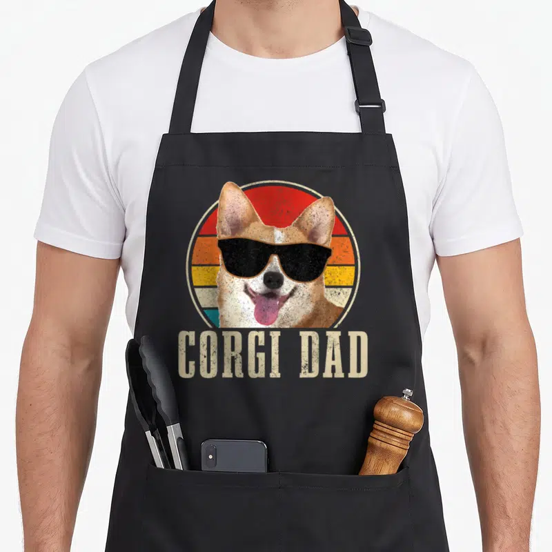 Corgi dog dad apron - Retro corgi with sunglasses in front of striped sun circle with text CORGI DAD below — shown on a black cotton canvas apron against a white background