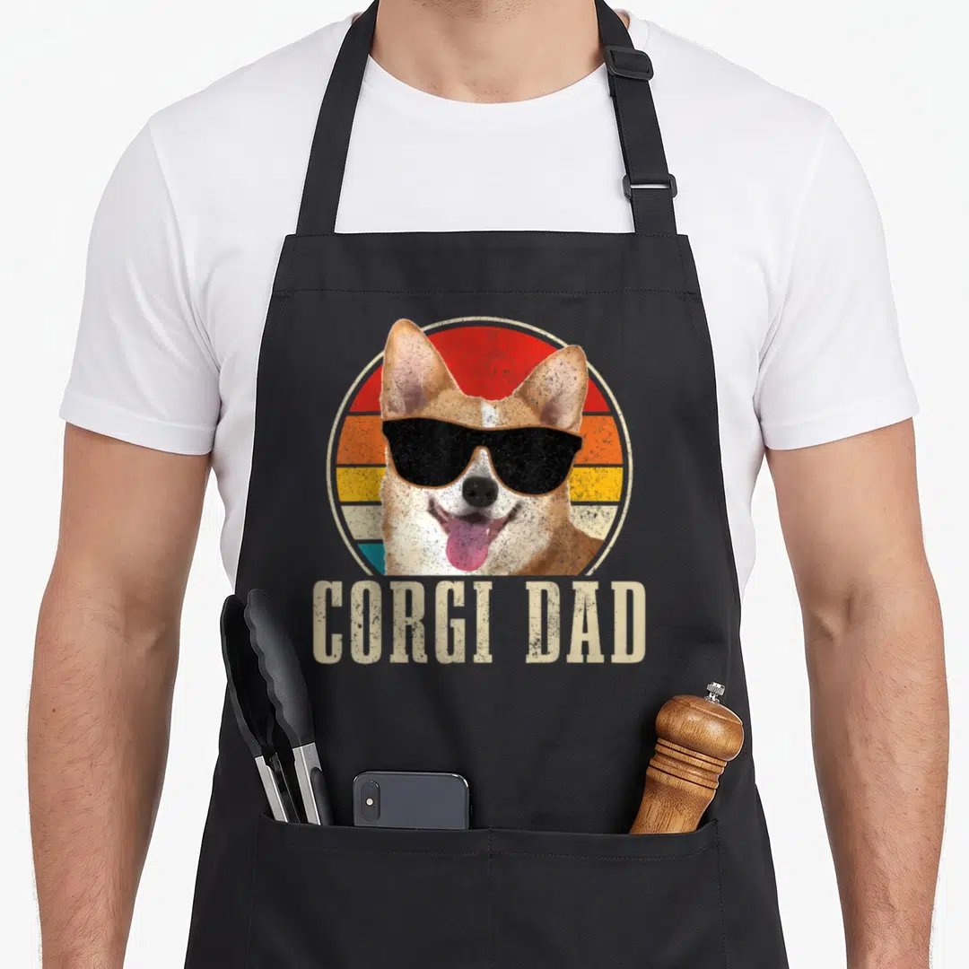 Corgi dog dad apron - Retro corgi with sunglasses in front of striped sun circle with text CORGI DAD below — shown on a black cotton canvas apron against a white background