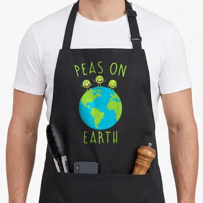 Earth peas apron - Three lime green peas standing on a blue and green globe under arched PEAS ON EARTH text — shown on a black cotton canvas apron against a white background