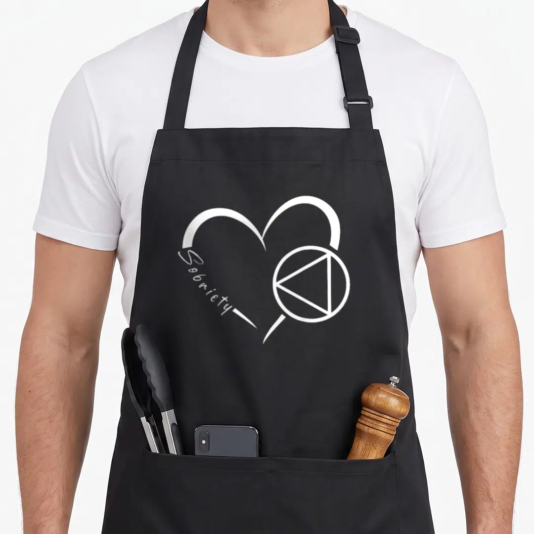 Alcoholic sobriety apron - White line art heart with Sobriety text and circle triangle sobriety symbol on black fabric — shown on a black cotton canvas apron against a white background