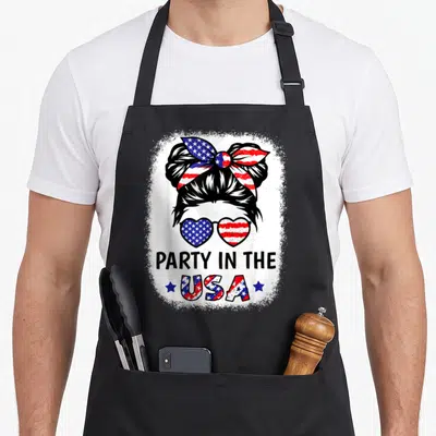 Cool flag 4th of july apron - Woman messy bun bow heart sunglasses flag pattern PARTY IN THE USA distressed print — shown on a black cotton canvas apron against a white background