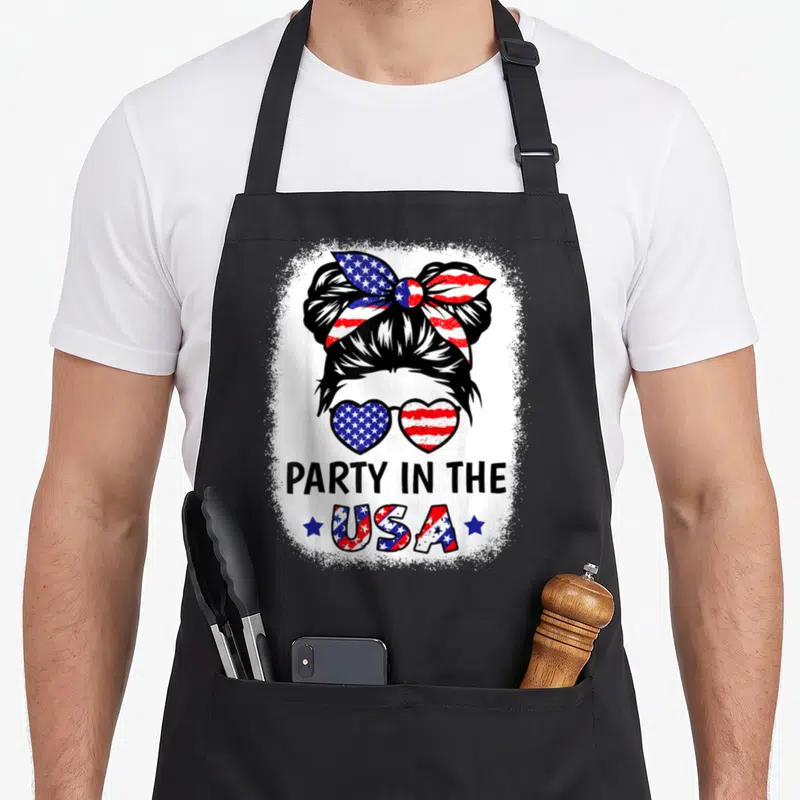Cool flag 4th of july apron - Woman messy bun bow heart sunglasses flag pattern PARTY IN THE USA distressed print — shown on a black cotton canvas apron against a white background