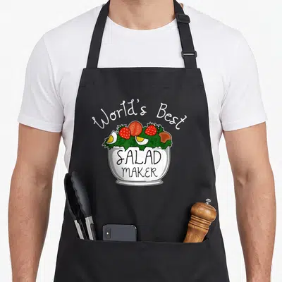 Salad veggie apron - Cartoon white bowl with red strawberries and yellow eggs labeled SALAD MAKER — shown on a black cotton canvas apron against a white background