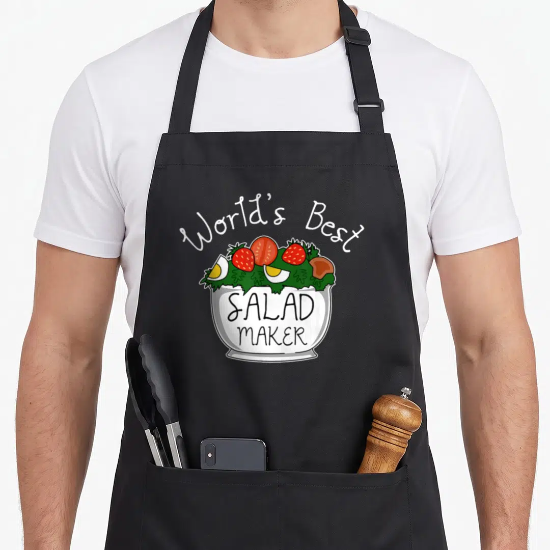 Salad veggie apron - Cartoon white bowl with red strawberries and yellow eggs labeled SALAD MAKER — shown on a black cotton canvas apron against a white background