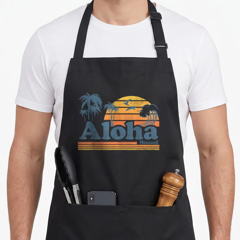 Aloha 70s apron - Retro sunset with blue palm trees, wave, birds, and Aloha Hawaii text in orange and yellow stripes — shown on a black cotton canvas apron against a white background
