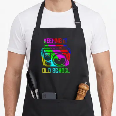 Keeping school 80s apron - Colorful striped boombox with KEEPING IT OLD SCHOOL text in retro font — shown on a black cotton canvas apron against a white background