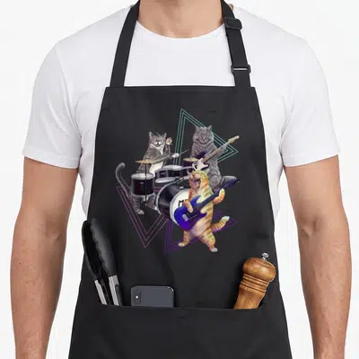 Band kitty cat apron - Three cartoon cats playing drums and guitars surrounded by neon pink and blue triangles — shown on a black cotton canvas apron against a white background