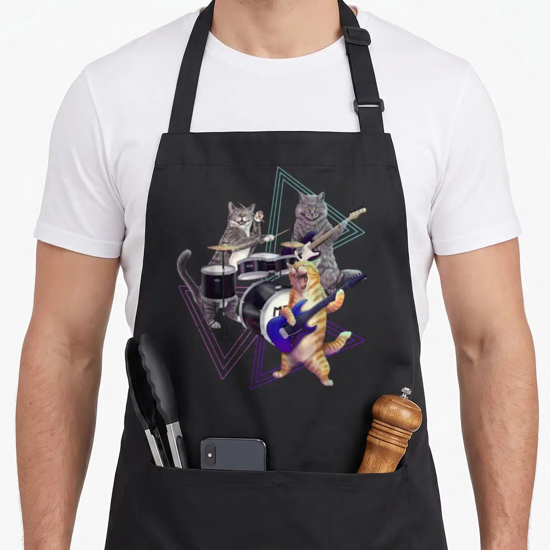 Band kitty cat apron - Three cartoon cats playing drums and guitars surrounded by neon pink and blue triangles — shown on a black cotton canvas apron against a white background