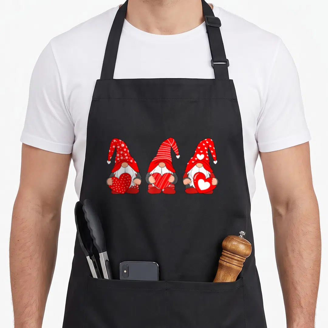 Three valentine's day apron - Three cartoon gnomes in a row holding red hearts with patterned hats — shown on a black cotton canvas apron against a white background
