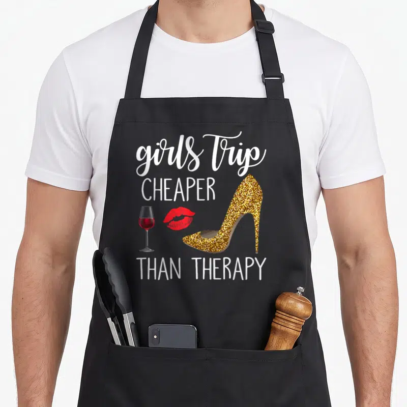 Weekend travel aprons - White text reading girls trip cheaper than therapy with gold heel, red lips, and wine glass — shown on a black cotton canvas apron against a white background