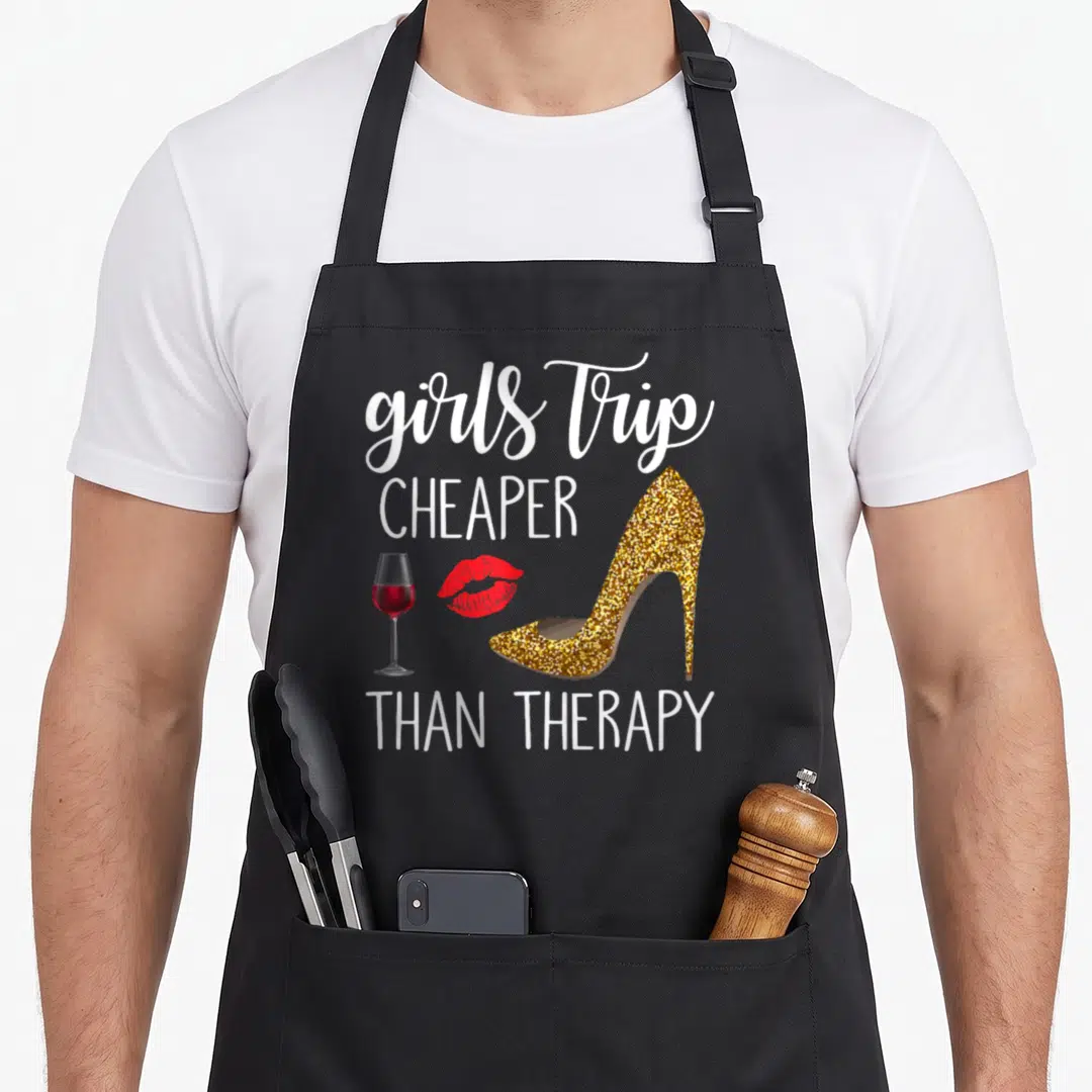 Weekend travel aprons - White text reading girls trip cheaper than therapy with gold heel, red lips, and wine glass — shown on a black cotton canvas apron against a white background