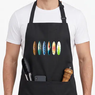 Sport summer aprons - Six cartoon surfboards in orange, teal, blue, cream, and green arranged horizontally — shown on a black cotton canvas apron against a white background