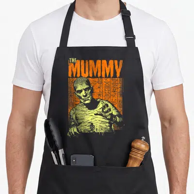 Universal mummy and me aprons - Distressed THE MUMMY text above a bandaged figure on orange and yellow stripes — shown on a black cotton canvas apron against a white background