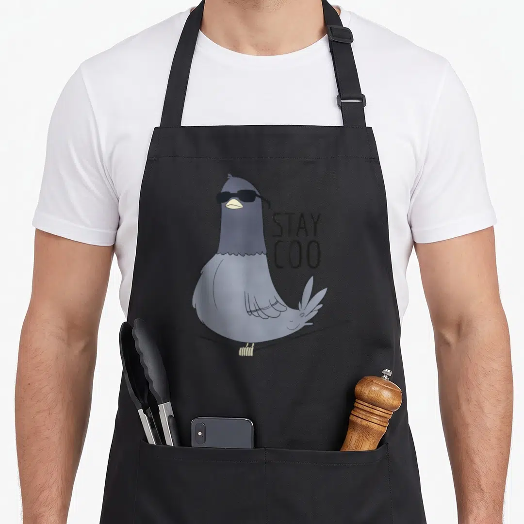 Coo bird apron - Cartoon blue-grey pigeon wearing black sunglasses on a wire next to stacked STAY COO text — shown on a black cotton canvas apron against a white background