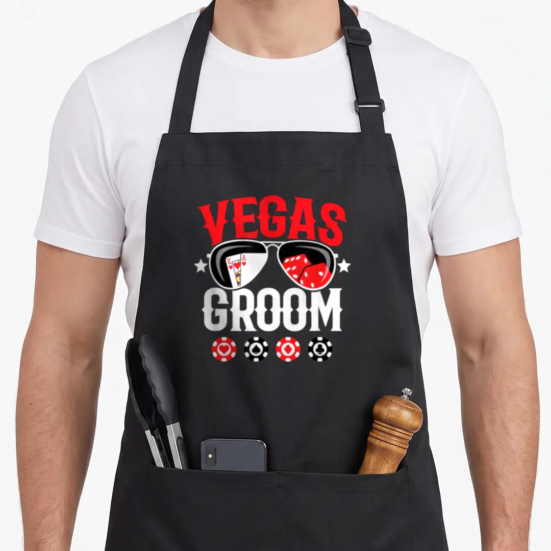 Bachelor vegas apron - VEGAS GROOM text over sunglasses reflecting cards dice and poker chips in red white black — shown on a black cotton canvas apron against a white background