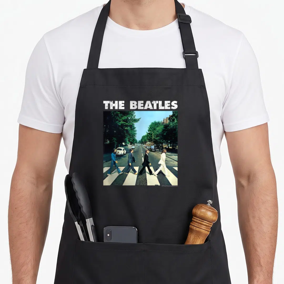 Cute abbey road apron - The Beatles text above four people walking on a crosswalk in blue and green — shown on a black cotton canvas apron against a white background