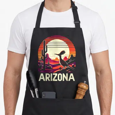 Roadrunner cactus apron - Retro Arizona sunset scene with roadrunner, cacti, mountains, and ARIZONA text in orange red — shown on a black cotton canvas apron against a white background