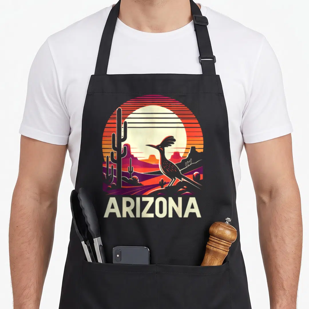 Roadrunner cactus apron - Retro Arizona sunset scene with roadrunner, cacti, mountains, and ARIZONA text in orange red — shown on a black cotton canvas apron against a white background