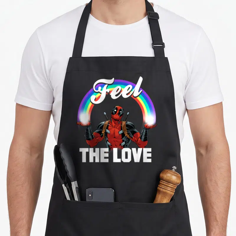 Feel rainbow apron - Deadpool character under arched Feel text and rainbow frame with THE LOVE below — shown on a black cotton canvas apron against a white background