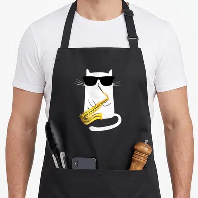 Saxophone cat wearing apron - White cartoon cat with black sunglasses playing a gold saxophone — shown on a black cotton canvas apron against a white background