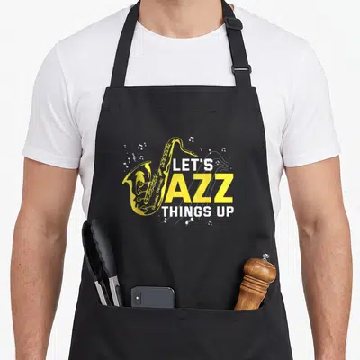 Saxophonist big band apron - Yellow cartoon saxophone overlapping white and yellow text Let's Jazz Things Up — shown on a black cotton canvas apron against a white background