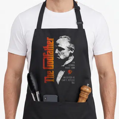 Vito fathers apron - Distressed white face with orange gradient text and red rose near chin — shown on a black cotton canvas apron against a white background