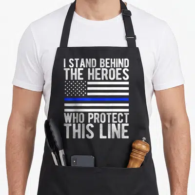 Thin line apron - White distressed text I STAND BEHIND HEROES around blue line flag on grey background — shown on a black cotton canvas apron against a white background