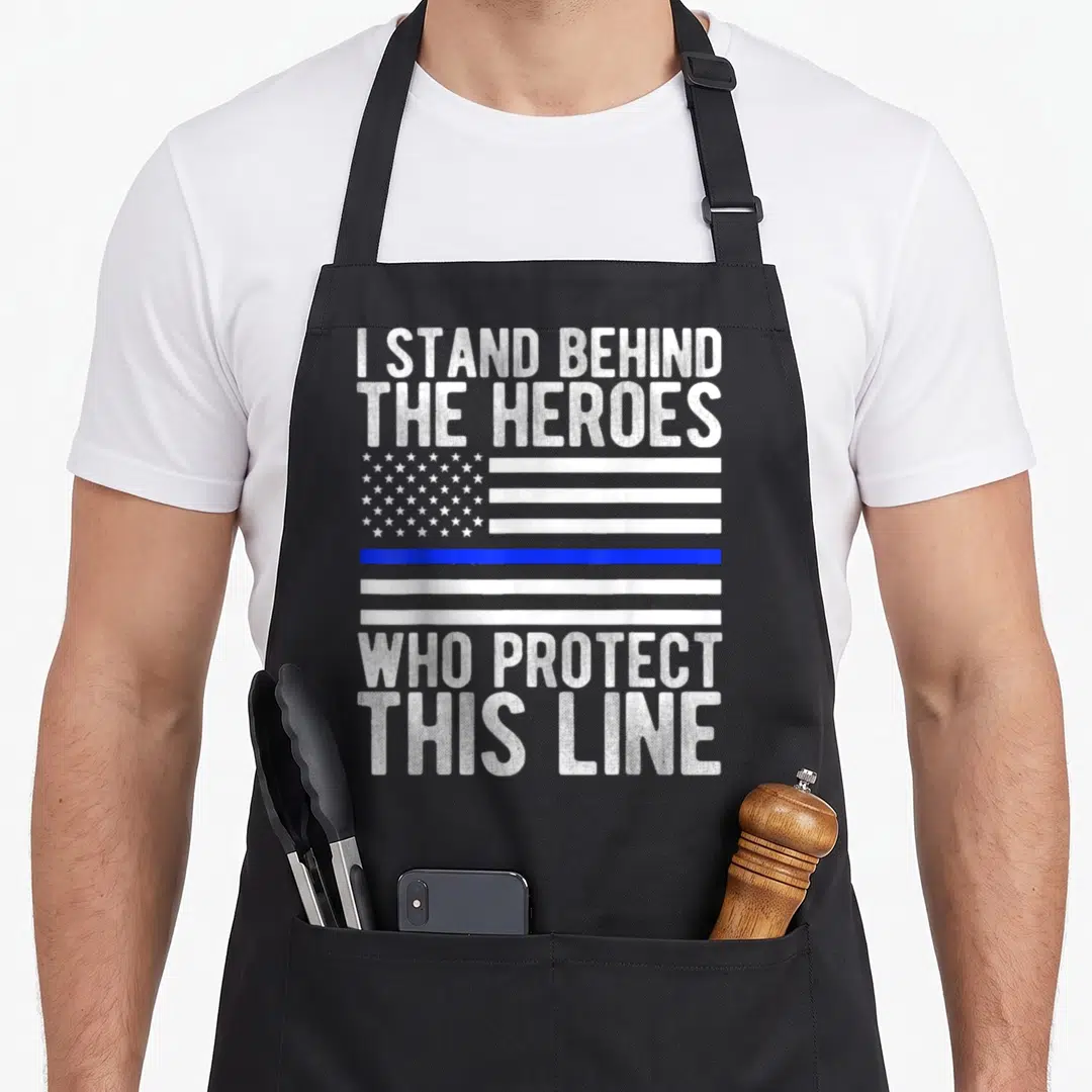 Thin line apron - White distressed text I STAND BEHIND HEROES around blue line flag on grey background — shown on a black cotton canvas apron against a white background
