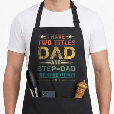 Title father day apron - Stacked multicolor distressed text reading I Have Two Titles Dad And Step-Dad I Rock Them Both — shown on a black cotton canvas apron against a white background