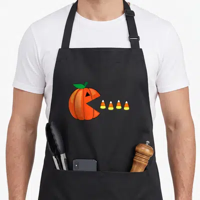 Corn halloween apron - Cartoon orange pumpkin with open mouth facing four candy corns in white yellow and orange — shown on a black cotton canvas apron against a white background