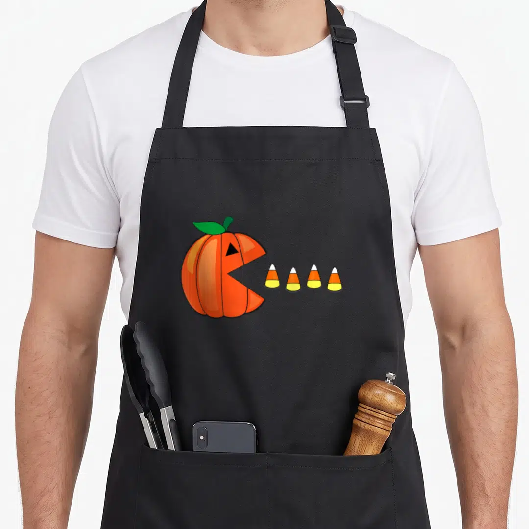 Corn halloween apron - Cartoon orange pumpkin with open mouth facing four candy corns in white yellow and orange — shown on a black cotton canvas apron against a white background