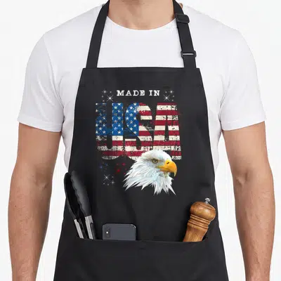 Patriotic bald american flag apron - MADE IN USA text with distressed flag pattern and bald eagle head in red white blue — shown on a black cotton canvas apron against a white background