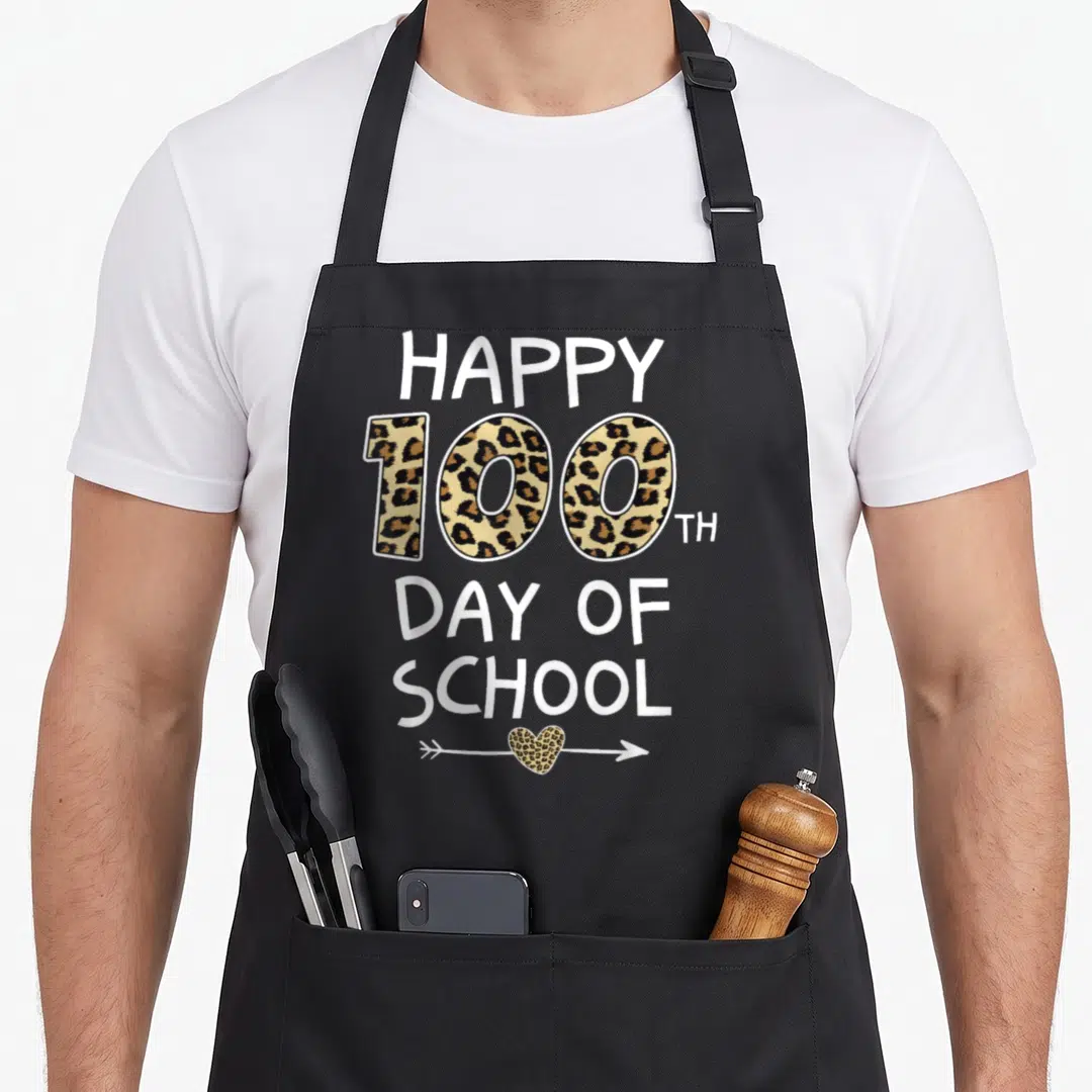 Leopard student teacher apron - Vertical Happy 100th Day of School text with leopard print 100 and arrow heart — shown on a black cotton canvas apron against a white background