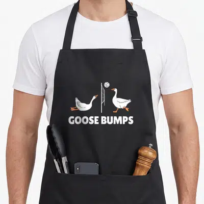 Volleyball goose apron - Two white geese playing volleyball with GOOSE BUMPS text below — shown on a black cotton canvas apron against a white background