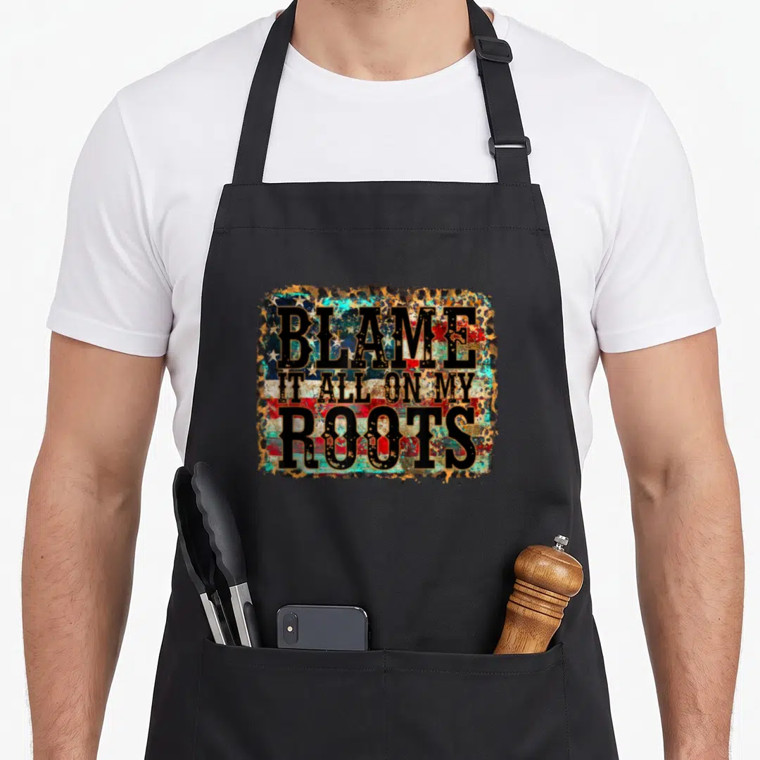 Blame cook's country aprons - Distressed text BLAME IT ALL ON MY ROOTS with flag and leopard fill inside rough border — shown on a black cotton canvas apron against a white background