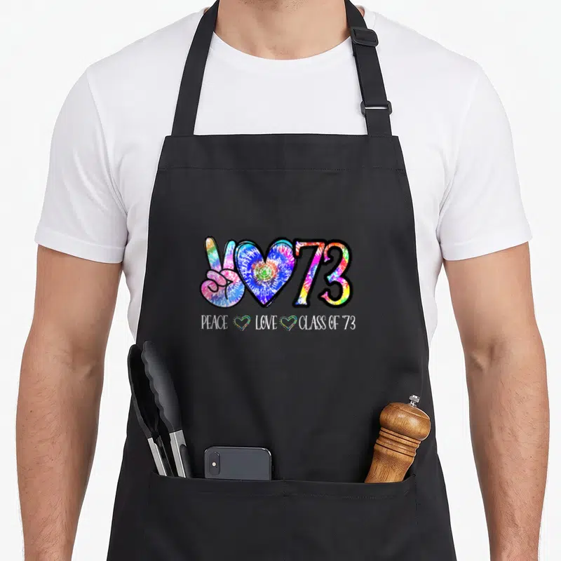 50th reunion tie-dye apron - Tie-dye peace hand, heart, 73 symbols with white PEACE LOVE CLASS OF '73 text and two — shown on a black cotton canvas apron against a white background
