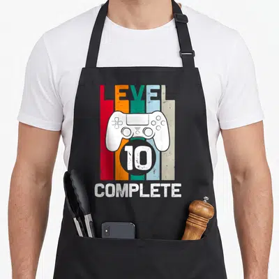Level wedding apron - Game controller graphic with LEVEL 10 COMPLETE text over vertical bars — shown on a black cotton canvas apron against a white background