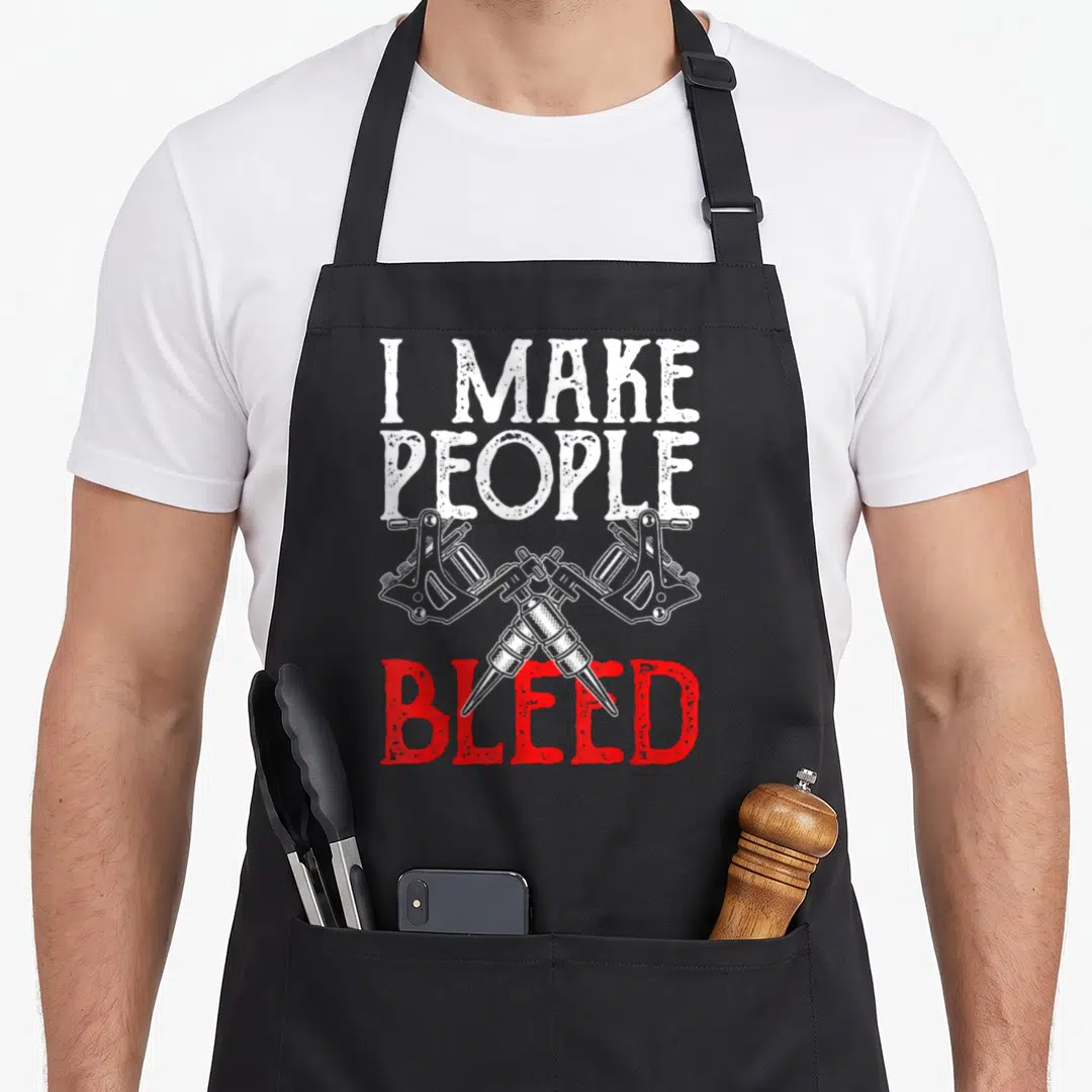 Master tattoo apron - Crossed tattoo machines with I MAKE PEOPLE BLEED text in white and red distressed font — shown on a black cotton canvas apron against a white background