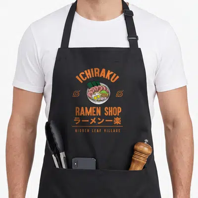Shippuden cartoon apron - Central ramen bowl graphic with orange ICHIRAKU text and Japanese characters flanked by swirl — shown on a black cotton canvas apron against a white background